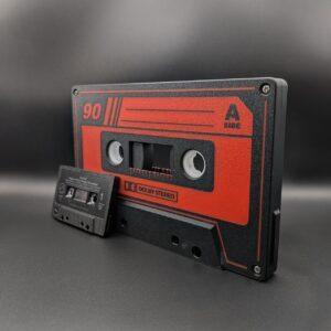 An Iconic Audio Cassette LED Nightlight in black and red, featuring a retro A Side and 90 design, stands upright like a vintage boombox centerpiece. Nearby, a smaller black cassette rests beside an artisanal turntable stylus on a dark surface against an acoustic-treated plain background.