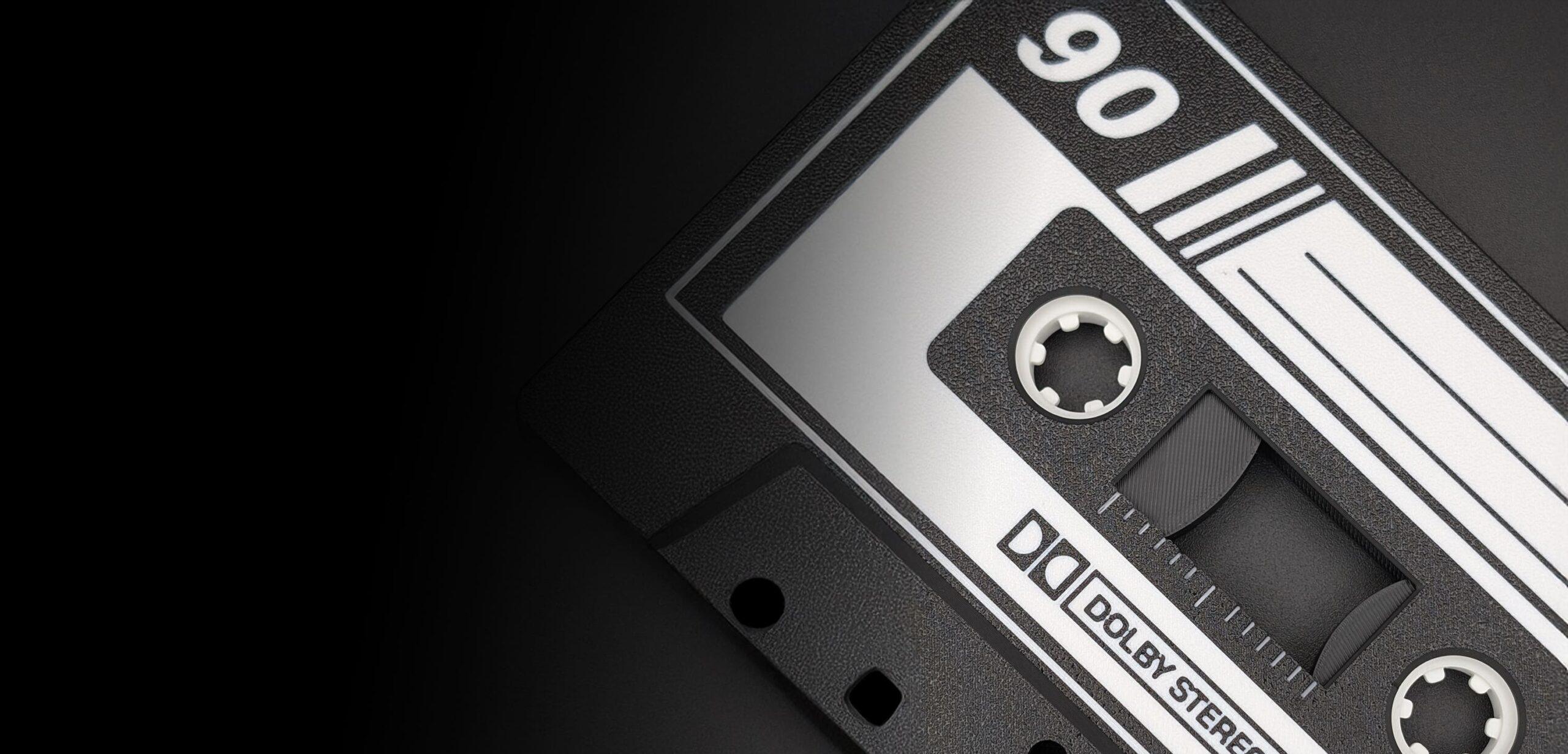Close-up of a vintage audio cassette tape featuring a black and white label, showcasing components like the magnetic tape reels, hub spools, and the precision-engineered capstan holes. The number 90 is prominently displayed on the shell casing, all set against a deep black background that accentuates the analog charm of this classic recording medium.