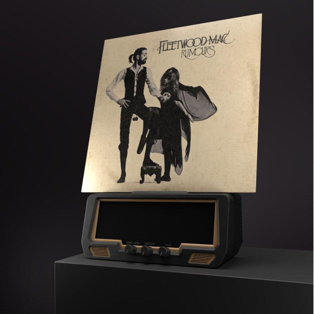 A Fleetwood Mac Rumours vinyl record is propped on a vintage radio, flanked by stereo speakers and audio cables, against a dark backdrop.