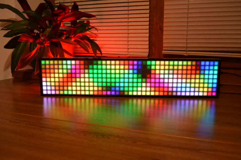A rectangular LED pixel display pulses a colorful, abstract pattern on a wooden floor by audio speakers, cables, and studio monitors.