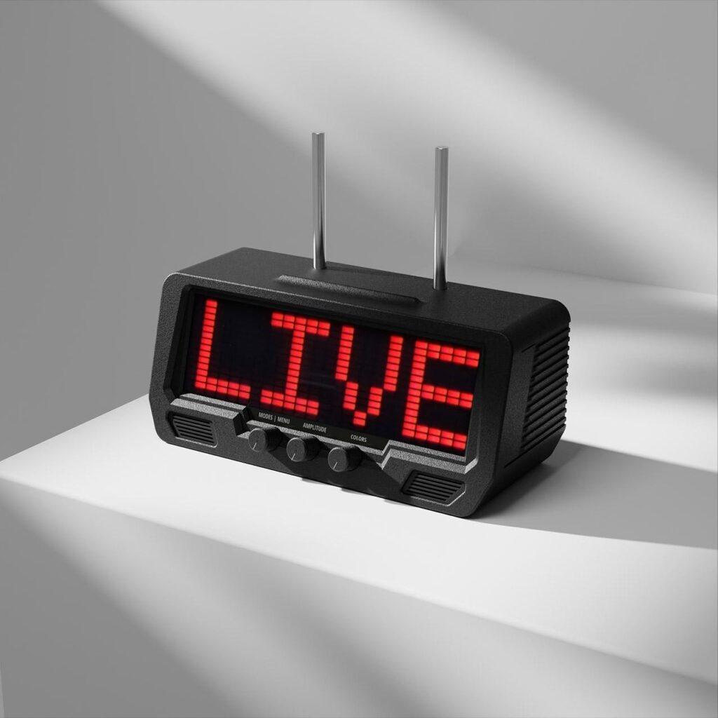 A digital clock radio with built-in speakers displays LIVE in red LED letters on its screen, resting on a white surface in bright lighting.
