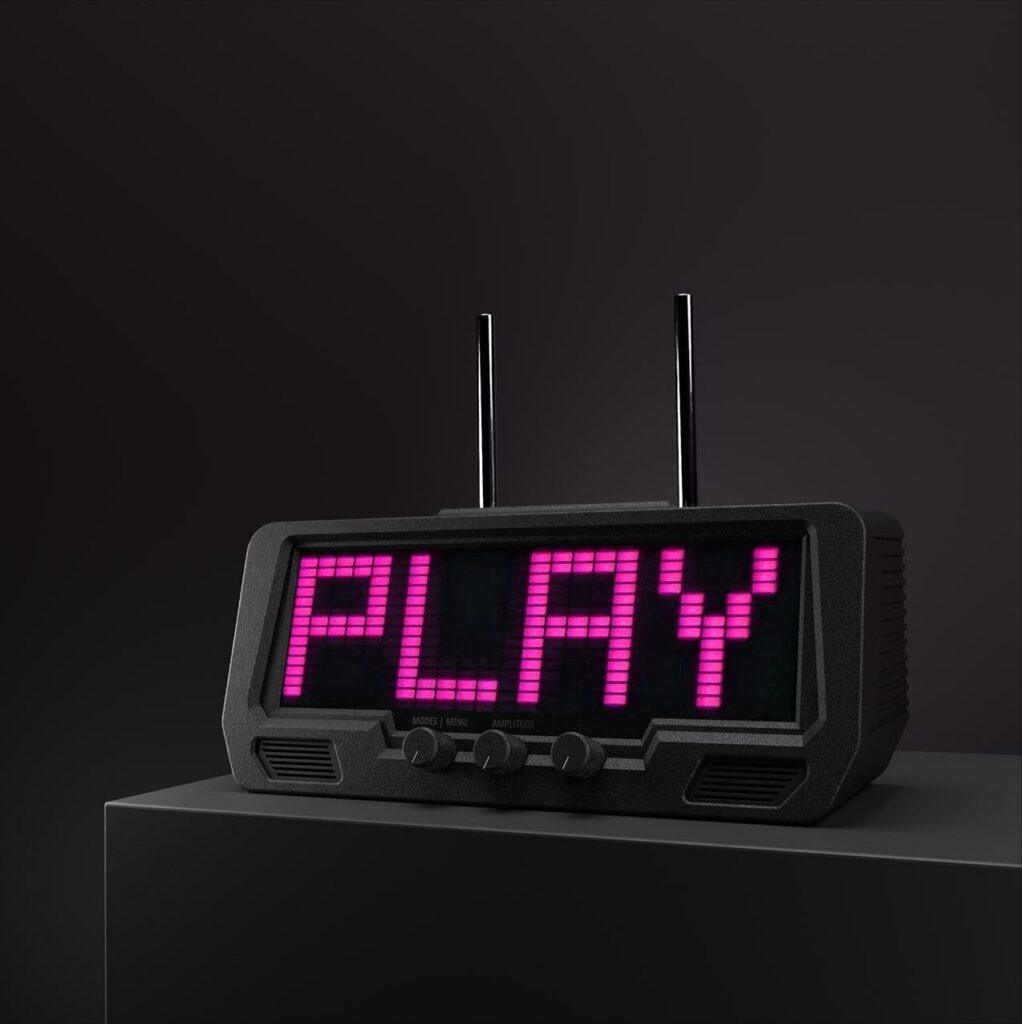 A matte-black stereo receiver with a digital pink LED display reading PLAY, sits on an audio rack against a shadowy background.